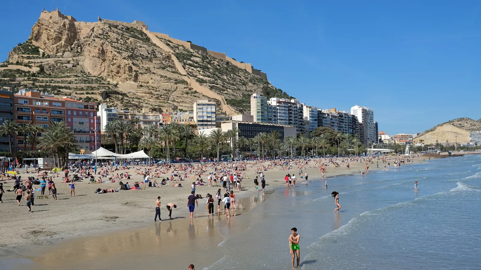 Kasteel Santa Bárbara op zijn rotsachtige heuvel boven het strand van Postiguet in Alicante, met zwemmers aan de rand van de zee en appartementsgebouwen langs de kust