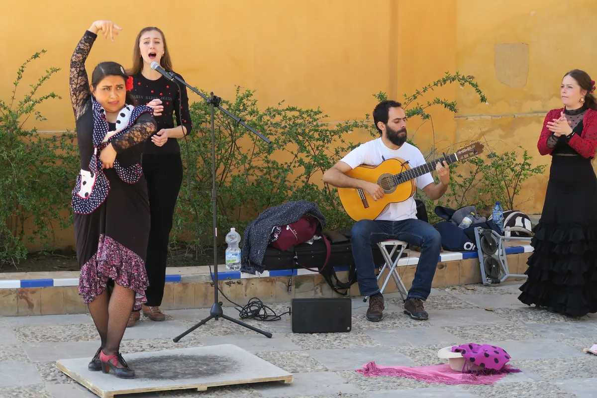 En flamencodanser i sjal med prikker, en sanger, en siddende guitarist og en klappende palmera optræder på en gård i Sevilla