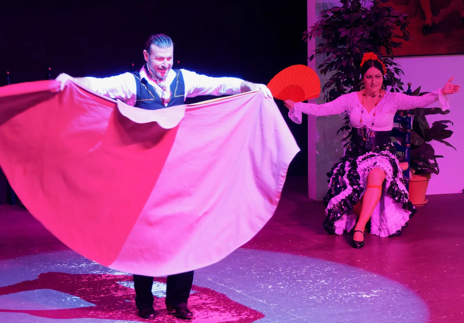 Een mannelijke danser in een vest spreidt een grote rood-witte cape wijd uit op een uitgelicht podium, terwijl een vrouwelijke danser in een zwarte jurk met stippen zit met een rode waaier omhoog, beiden vastgelegd midden in hun optreden